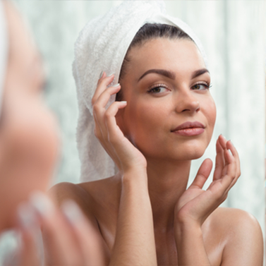 A woman with a towel on her head applies skincare in front of a mirror.